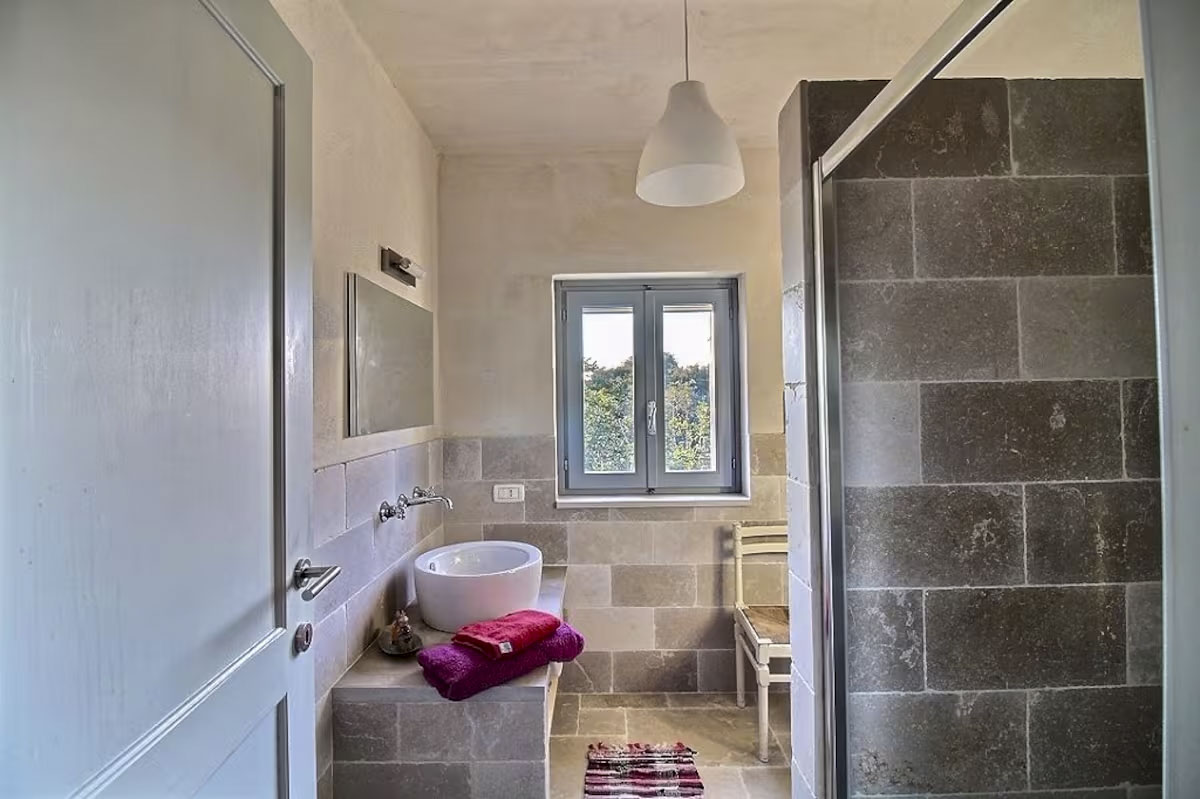 Modern bathroom with stone-tiled shower, vessel sink, and light grey window frame.