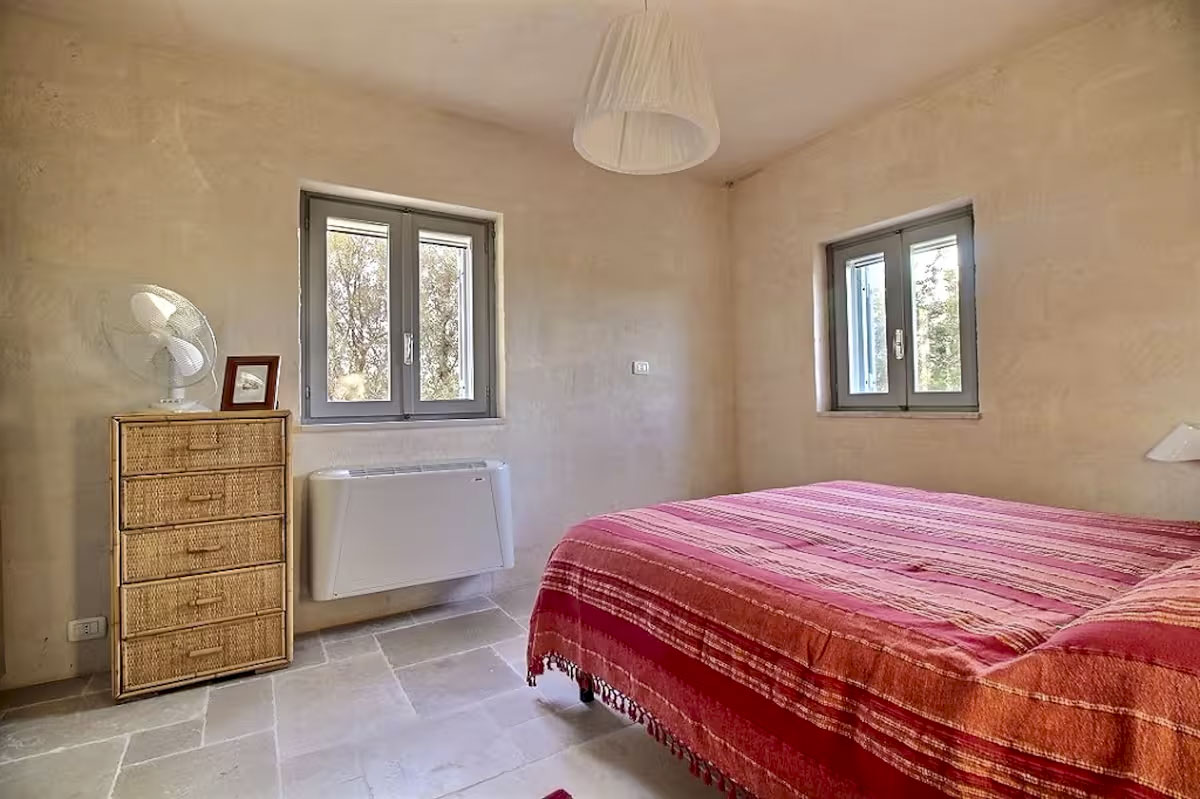 Cozy main bedroom with red-striped duvet, rattan dresser, stone Chianca floor, and two windows facing the exterior.