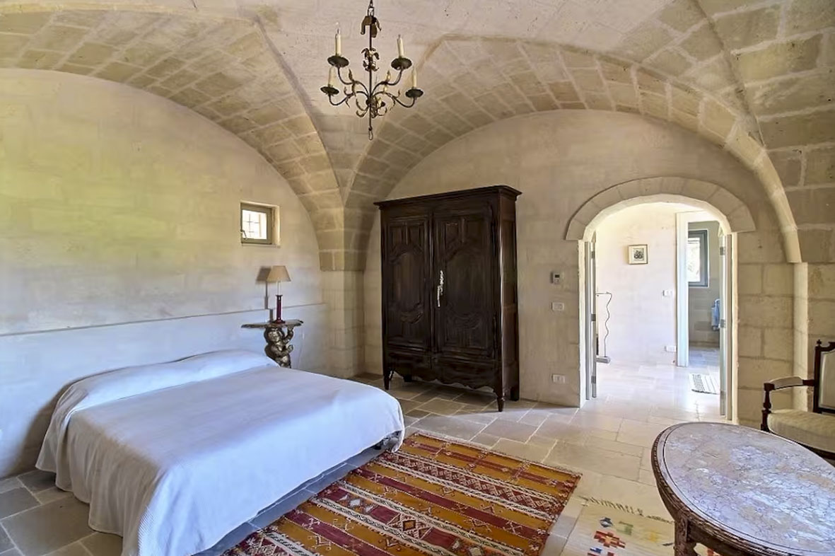 Spacious bedroom in Agriturismo Masseria apartment with stone vaulted ceiling, antique wardrobe, and view into the bathroom.